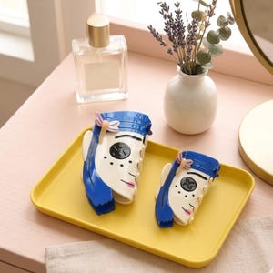 May include: Two decorative hair clips in the shape of a stylized face with blue hair, white skin, and pink lips. They are placed on a yellow rectangular tray. A perfume bottle and a small vase with flowers are in the background.