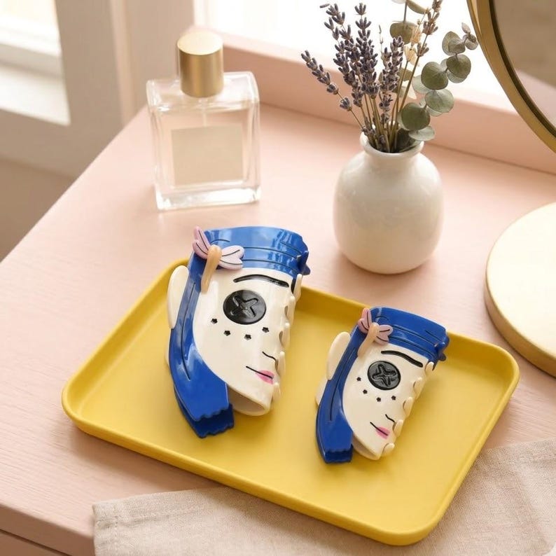 May include: Two decorative hair clips in the shape of a stylized face with blue hair, white skin, and pink lips. They are placed on a yellow rectangular tray. A perfume bottle and a small vase with flowers are in the background.