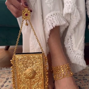 Op de afbeelding: Een rechthoekige goudkleurige metalen clutch met gedetailleerde bloemmotieven in reliëf, een centraal cirkelvormig motief en kralenfranjes langs de randen. De tas heeft een goudkleurig kettinghengsel met een vierkant accentstuk met een rode steen.