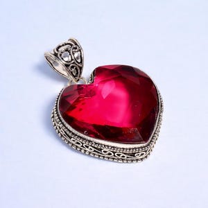 May include: A heart-shaped pendant featuring a large, faceted, deep red gemstone. The gemstone is set in a silver-coloured metal frame with an ornate design. The pendant has a decorative bail for attaching to a chain.