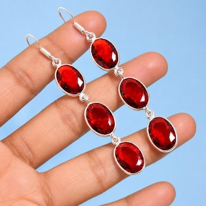 Puede incluir: Un par de pendientes largos colgantes con tres piedras preciosas ovaladas de color rojo intenso facetadas, engastadas en metal plateado pulido. Cada pendiente consta de una fila vertical de piedras unidas por pequeños eslabones plateados.