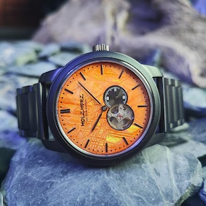 Op de afbeelding: Een zwart polshorloge met een oranje wijzerplaat en een zwarte metalen band. De wijzerplaat heeft de tekst "HOLZ HERZ" en "METEOR EDITION". Het horloge heeft een kleine subwijzerplaat en een zichtbaar mechanisch uurwerk. Het horloge ligt op grijze rotsen.