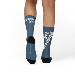 May include: A pair of blue socks with black heels and toes. The text "see ya!" is printed on one sock, and "get dropped" is printed on the other. The words "Speedy Cyclist Co." are printed on the side of one sock.