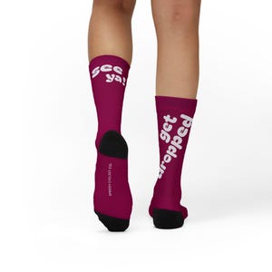 May include: Burgundy socks with black heels and toes. The left sock features the words "see ya!" in white, and the right sock has the words "get dropped" in white. The words "Speedy Cyclist Co." are printed on the side of the left sock.