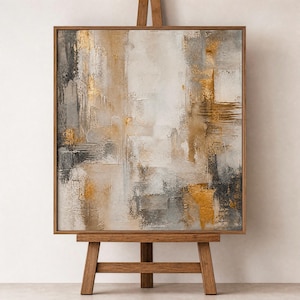 May include: Abstract art painting in a wooden frame. The artwork features a textured surface with a palette of white, gray, gold, and brown. The painting is displayed on a wooden easel, showcasing the artwork's colors and textures.