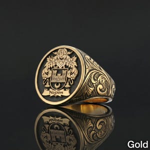 May include: A gold signet ring with an engraved design. The ring features a crest with a castle and the name "Negron" on a banner. The band of the ring has an ornate, swirling pattern. The word "Gold" is in the bottom right corner.