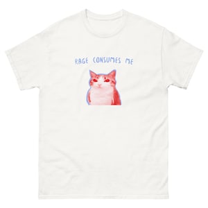 Puede incluir: Camiseta blanca con un gráfico de gato rojo y blanco con contorno azul. El texto "RAGE CONSUMES ME" está impreso en azul encima del gato. La camiseta es de manga corta y cuello redondo.