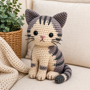 Peut inclure: Poupée chat au crochet faite à la main. Le chat est beige avec des rayures grises, des yeux noirs et un nez rose. Il est assis, avec une longue queue et des moustaches noires. La poupée est posée sur une surface beige.