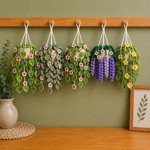 May include: Five handcrafted crochet hanging planters with white pots and detailed floral and leaf patterns. The planters are suspended from a wooden beam, displaying a range of colours including green, pink, yellow, and purple. A framed botanical print rests on a wooden surface.