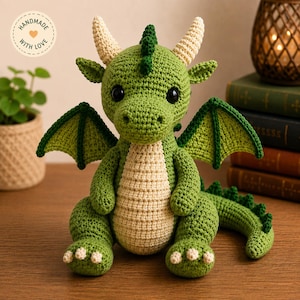 May include: A handmade crochet dragon in green and cream. The dragon has large black eyes, cream horns, and green wings. The dragon is sitting on a wooden surface. The image includes a small plant and a candle. The text "Handmade with Love" is visible.
