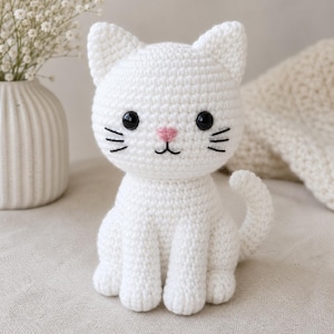 May include: A handmade, white crochet cat figurine with black eyes, whiskers, and a pink nose. The cat is sitting upright with its tail curved to the side. The cat is a soft, plush toy.