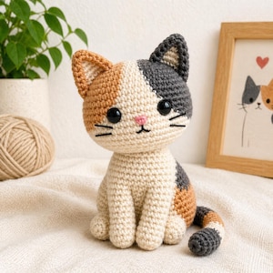 Può includere: Un gatto all'uncinetto fatto a mano in crema, marrone e grigio. Il gatto amigurumi ha grandi occhi neri, un naso rosa e una coda a strisce. Un gomitolo di lana e un'immagine incorniciata di due gatti sono sullo sfondo.