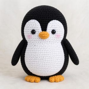 May include: A handmade crochet penguin with a black head, wings, and body outline. The penguin has a white belly and face, with a yellow beak and feet. The penguin has small black eyes and pink cheeks. This soft toy is perfect for children.