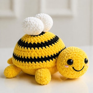 May include: A crocheted bee toy with a yellow body, black stripes, and white pom-pom eyes. The bee has a smiling face with rosy cheeks and small, yellow legs. The toy is made of soft, textured yarn.