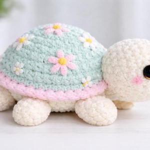 Può includere: Una tartaruga all'uncinetto fatta a mano. Il guscio è verde chiaro, decorato con fiori rosa e bianchi, bordato di rosa. La testa e le zampe sono bianche, con occhi neri e guance rosa. Un giocattolo carino.