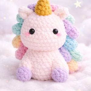 May include: A handmade crochet unicorn plush toy. The unicorn has a light pink body, a yellow horn, and a rainbow mane. It has black eyes and pink cheeks. The plush is sitting on a white, fluffy surface.