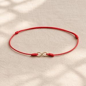 May include: A thin red cord bracelet featuring a central gold-toned infinity symbol charm. The bracelet is secured with adjustable sliding knots on either side of the charm and two additional knots along the cord to maintain its circular shape.