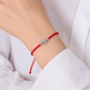 Puede incluir: Una pulsera de cordón rojo con un dije de flor plateado central flanqueado por dos pequeñas cuentas plateadas. La pulsera está construida con un diseño de cordón trenzado e incluye extremos colgantes ajustables para un ajuste seguro.