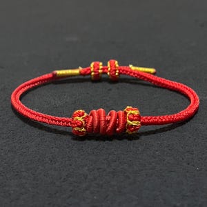 May include: A red cord bracelet featuring a central decorative knot element with gold-coloured thread accents. The bracelet includes two smaller sliding knots for adjustment and two gold-coloured metal tips at the ends of the cord.