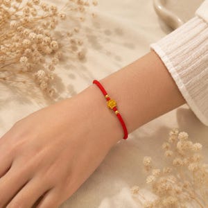 May include: A thin red cord bracelet featuring a small, textured gold-colored flower charm at the center, flanked by two tiny gold-colored beads. The bracelet is secured with a simple knot closure, creating a delicate and minimalist wrist accessory.