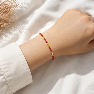 Op de afbeelding: Een dunne rode koordarmband met drie kleine, cilindervormige goudkleurige metalen kralen die gelijkmatig over de lengte van het koord zijn verdeeld. De armband wordt om de pols gedragen en toont de levendige rode kleur van het koord en de metalen afwerking.