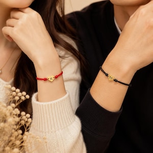 Pode incluir: Duas pulseiras de cordão ajustável com pequenos pingentes de coração em tom dourado. Uma pulseira tem um cordão vermelho com a letra K gravada no coração, e a outra um cordão preto com a letra M gravada no coração, ambas nos pulsos.