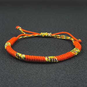 May include: A handmade orange bracelet with adjustable cords. The bracelet features a woven design with accents of yellow, green, and brown. The bracelet is a simple, yet stylish accessory.