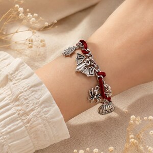 Könnte beinhalten: Ein Bettelarmband mit silberfarbenen Metallanhängern in Form einer Fledermaus, einer Spinne, eines Spinnennetzes und eines Kürbisses. Das Armband besteht aus einer silberfarbenen Metallkette, die mit einem dunkelroten Samtband verflochten ist.