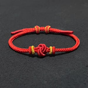 Könnte beinhalten: Ein leuchtend rotes Kordelarmband mit einem zentralen dekorativen Knoten, der von zwei kleinen goldfarbenen Metallzylindern flankiert wird. Das Armband verfügt über einen verstellbaren Schiebeverschluss mit zwei weiteren goldenen Akzenten an den Enden.