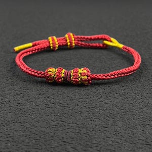 May include: Red and gold adjustable bracelet. The bracelet features a woven red cord with gold accents and decorative knots. The ends of the cord are finished with gold-coloured tips. The bracelet is designed to be adjustable for a comfortable fit.