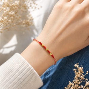 May include: A thin red braided cord bracelet featuring four small, polished gold-toned spherical beads spaced evenly along the centre. The bracelet is constructed from a fine, woven red thread that creates a delicate and minimalist appearance on the wrist.