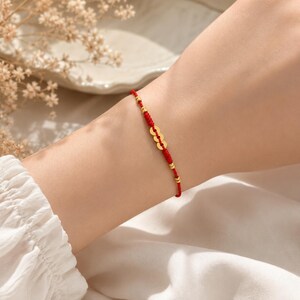 May include: A thin red cord bracelet featuring three small, circular gold-toned metal charms arranged in the center, accented by four tiny gold-toned beads spaced along the cord, creating a delicate and minimalist wrist accessory.