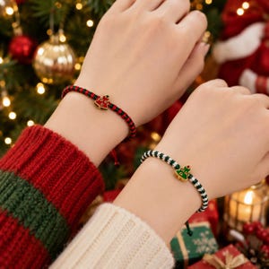 Könnte beinhalten: Zwei geflochtene Armbänder mit kleinen Metallanhängern in Form von Weihnachtsbäumen. Ein Armband hat eine rot-grüne Kordel mit einem roten Emaille-Baum, das andere eine weiß-grüne Kordel mit einem grünen Emaille-Baum.