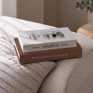 Könnte beinhalten: Zwei rechteckige Dekobücher, die übereinander gestapelt sind. Das obere Buch ist weiß mit dem Text "timeless SIMPLE living" und einem kleinen Foto. Das untere Buch ist braun mit dem Text "NATURAL SPACES" in weißen Großbuchstaben.