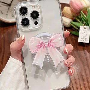 May include: Clear phone case with a pink bow-shaped grip. The case has a clear back and a clear bumper. The bow is light pink with white accents. The phone case is held in a hand.