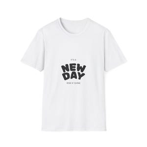 Può includere: T-shirt bianca con la scritta "IT'S A NEW DAY RISE N' SHINE" in nero, in grassetto e con carattere arrotondato. La maglietta ha maniche corte e girocollo. Il testo è centrato sul davanti.