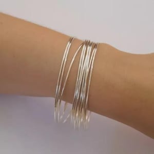 May include: A collection of thin, silver bangles stacked on a wrist. The bangles are made of a shiny, reflective material and appear lightweight. The bracelets are simple and elegant, suitable for everyday wear.