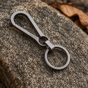 Può includere: Un portachiavi a moschettone in metallo grigio opaco dotato di un meccanismo di chiusura a molla con cinque linee orizzontali incise sul lato, collegato a un anello circolare per le chiavi, realizzato in un unico pezzo di metallo.