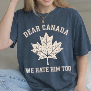 May include: A dark blue short-sleeve t-shirt featuring a large, distressed-style cream-colored maple leaf graphic in the center. The text "DEAR CANADA" is printed in a cream-colored arched font above the leaf, and "WE HATE HIM TOO" below.