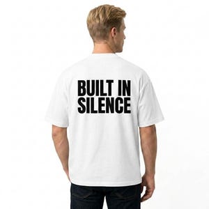 May include: A white t-shirt with the text "BUILT IN SILENCE" printed in black. The shirt is being worn by a person from the back, with the text centered on the upper back. The person is wearing dark blue jeans.