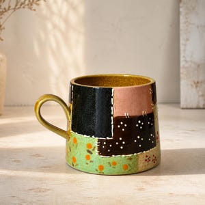 May include: A ceramic mug featuring a patchwork design with blocks of black, dusty pink, and dark brown with white dots. The lower section is light green with small orange and red floral patterns. It has a curved handle and a mustard yellow interior.