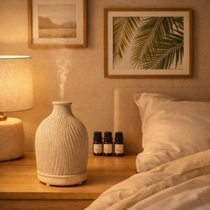Op de afbeelding: Een beige keramische diffuser voor etherische olie met een textuur van golvende verticale lijnen, die een dunne stroom mist uitstoot. De diffuser heeft een ronde, vaasachtige vorm en een kleine aan-uitknop aan de onderkant bij drie flesjes.