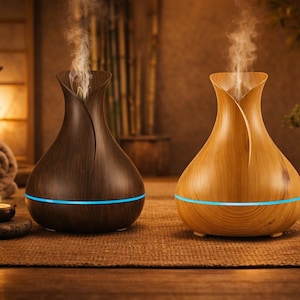 May include: Two aromatherapy diffusers, one dark brown and one light brown, with a wood grain finish. Both have a blue light band and emit a vapor. The scene includes a lit candle, stones, and a bonsai tree, creating a spa-like ambiance.