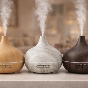 May include: Three essential oil diffusers in a row, each with a different wood grain finish. The diffusers are light brown, white, and dark brown. Each diffuser has a small control panel and is emitting a plume of vapor.