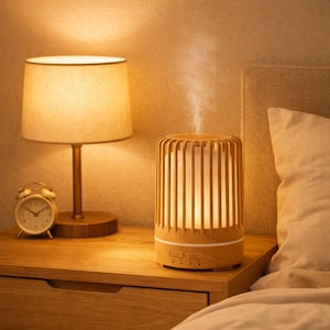 Könnte beinhalten: Ein zylindrischer Diffusor für ätherische Öle in heller Holzoptik mit einem lamellierten Außengehäuse und einem weißen Innenkern, der einen feinen Nebel abgibt. Das Gerät verfügt über ein horizontales Bedienfeld mit Tasten an der Basis.