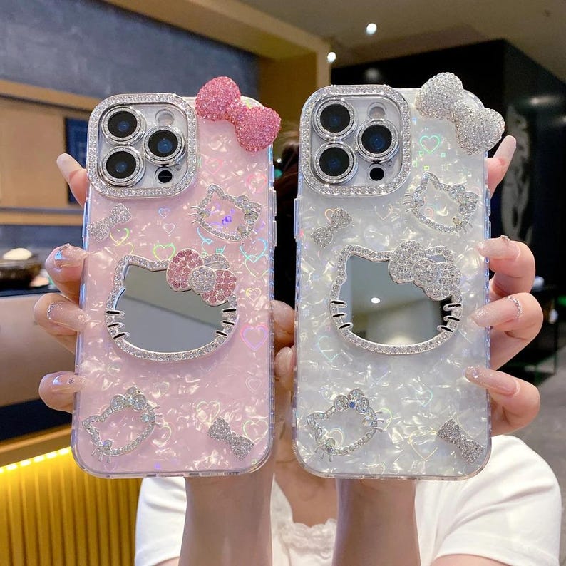 May include: Two phone cases, one pink and one white, each adorned with a Hello Kitty design. The cases feature sparkling rhinestones around the camera and the Hello Kitty face, and a bow on top. The pink case has a pink bow, while the white case has a white bow.