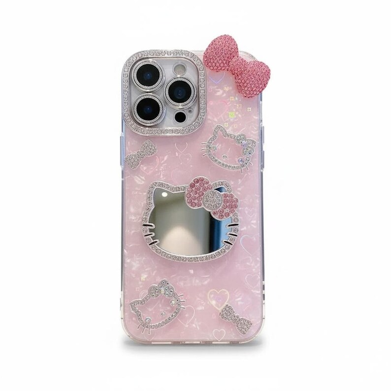 May include: Pink phone case with a glittery finish, featuring a Hello Kitty design. The case has a mirror, rhinestone accents, and a 3D bow. The camera lens is surrounded by rhinestones. The case is designed for a smartphone.