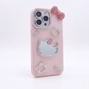 May include: Pink phone case with a bow and Hello Kitty embellishments. The case features a large Hello Kitty face with a bow, and several smaller bow and Hello Kitty designs. The camera lenses are surrounded by a silver border.