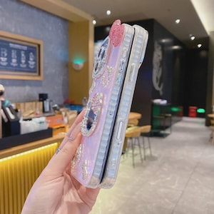 May include: A pink and clear phone case with a bow and jewel embellishments. The case has a glittery, iridescent finish and is decorated with rhinestones and mirrored accents. The phone case is held in a person's hand.