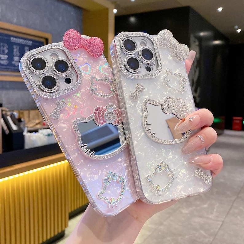 May include: Two decorative phone cases, one pink and one white, adorned with sparkling rhinestones and bow accents. The cases feature a mirrored Hello Kitty design and are embellished with additional crystal details.